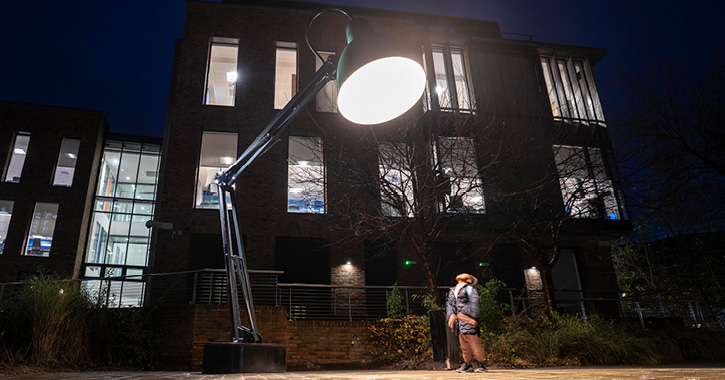 little boy stands directly under the light of Lumiere artwork Lampounette, which looks like a giant office desk lamp